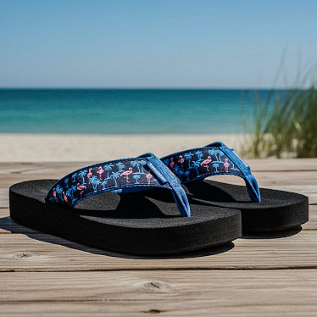 Pair of black sandals with blue straps featuring pink flamingos on a wooden surface with a beach background.