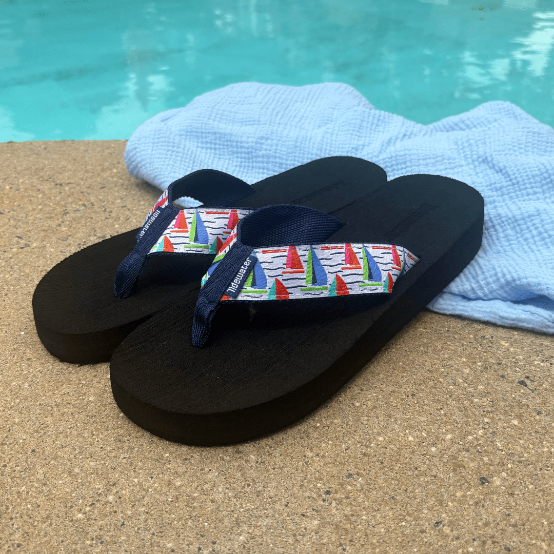 Black flip-flops with a colorful sailboat design on the straps, resting on a sandy surface near a pool.