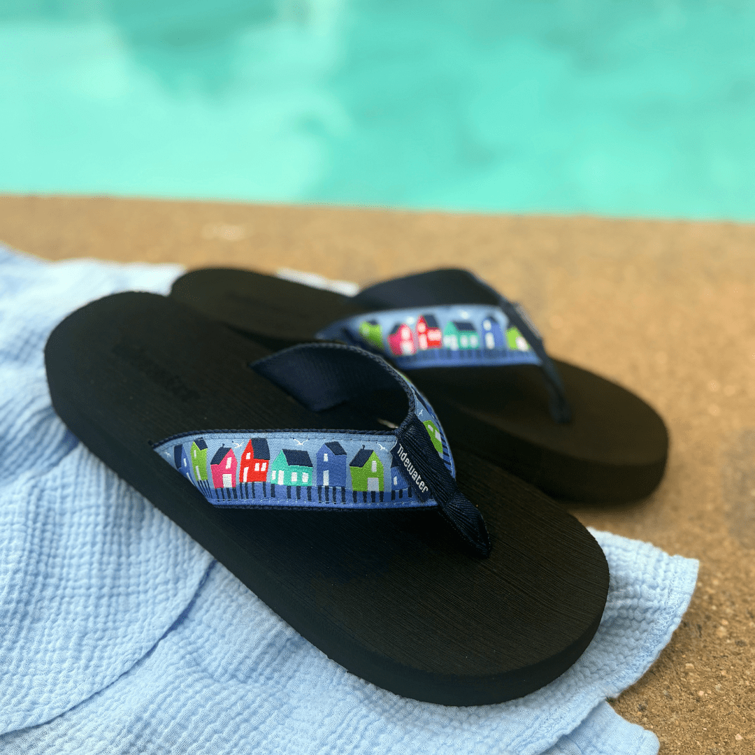 Navy flip-flops with colorful house designs on the straps, resting on a sandy surface near a pool.