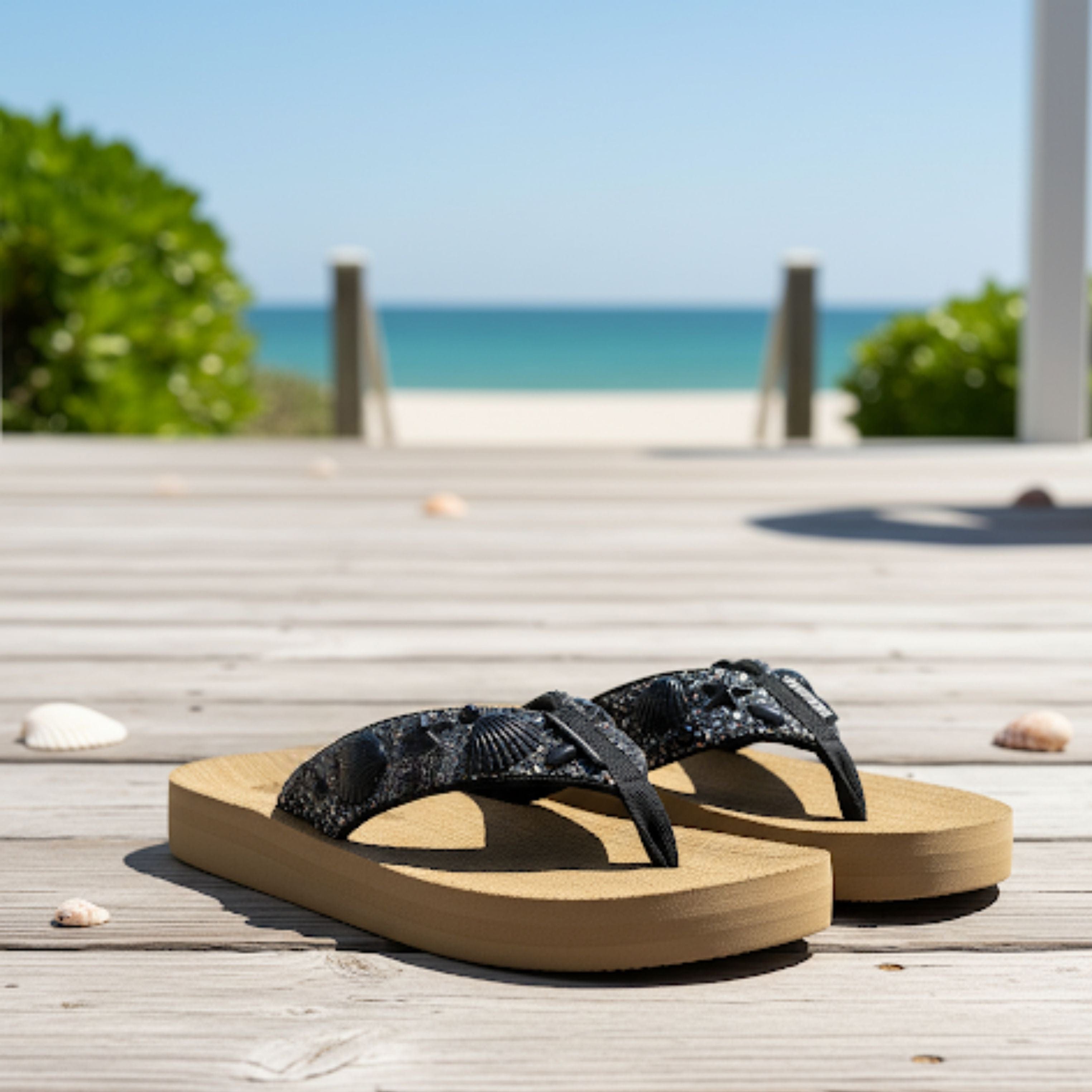 Black flip-flops with a textured strap on a wooden deck, overlooking a sunny beach and ocean. Perfect for summer outings.