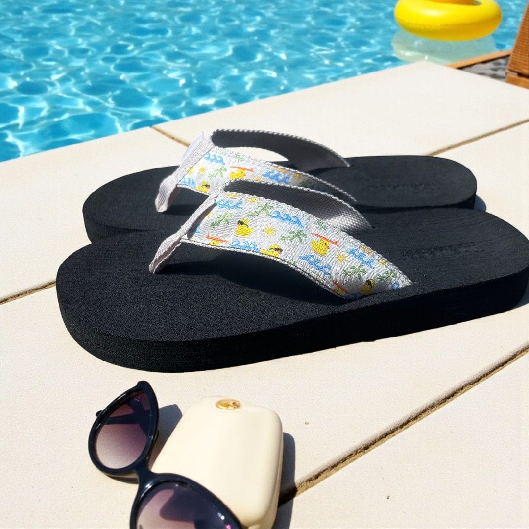 Black flip-flops with a colorful beach-themed strap resting on a poolside deck, next to sunglasses and a sunhat.