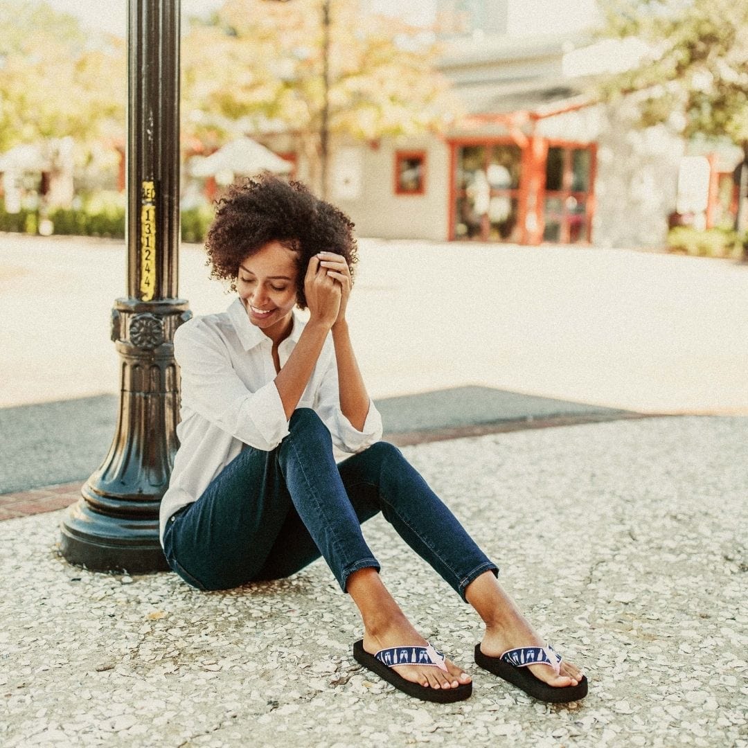A woman sits on the ground near a lamp post, wearing Rose All Day sandals, a white shirt, and dark jeans, smiling.