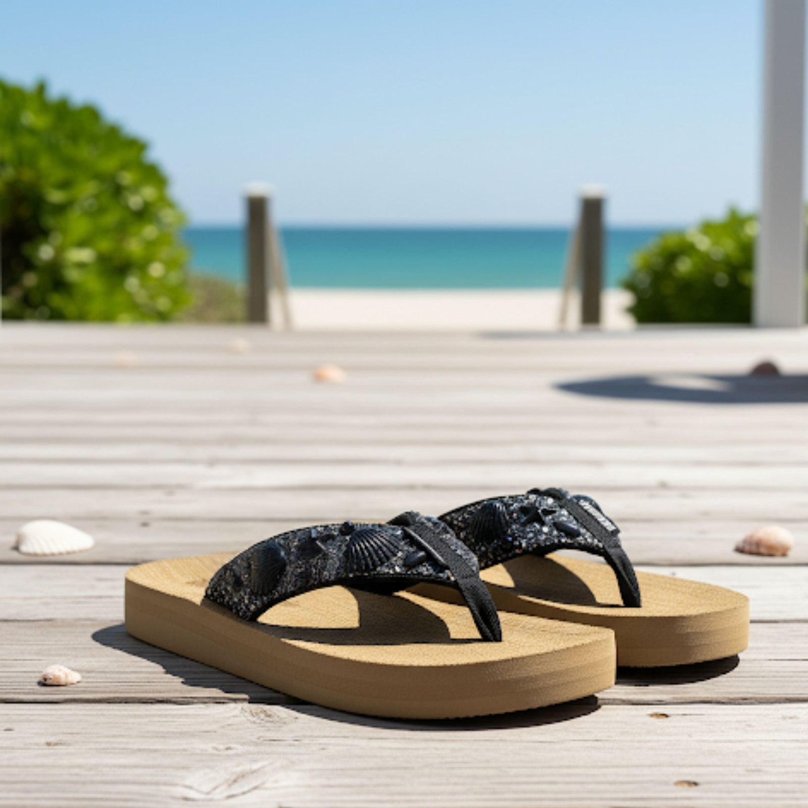 Black flip-flops with a textured strap on a wooden deck, overlooking a sunny beach and ocean. Perfect for summer outings.
