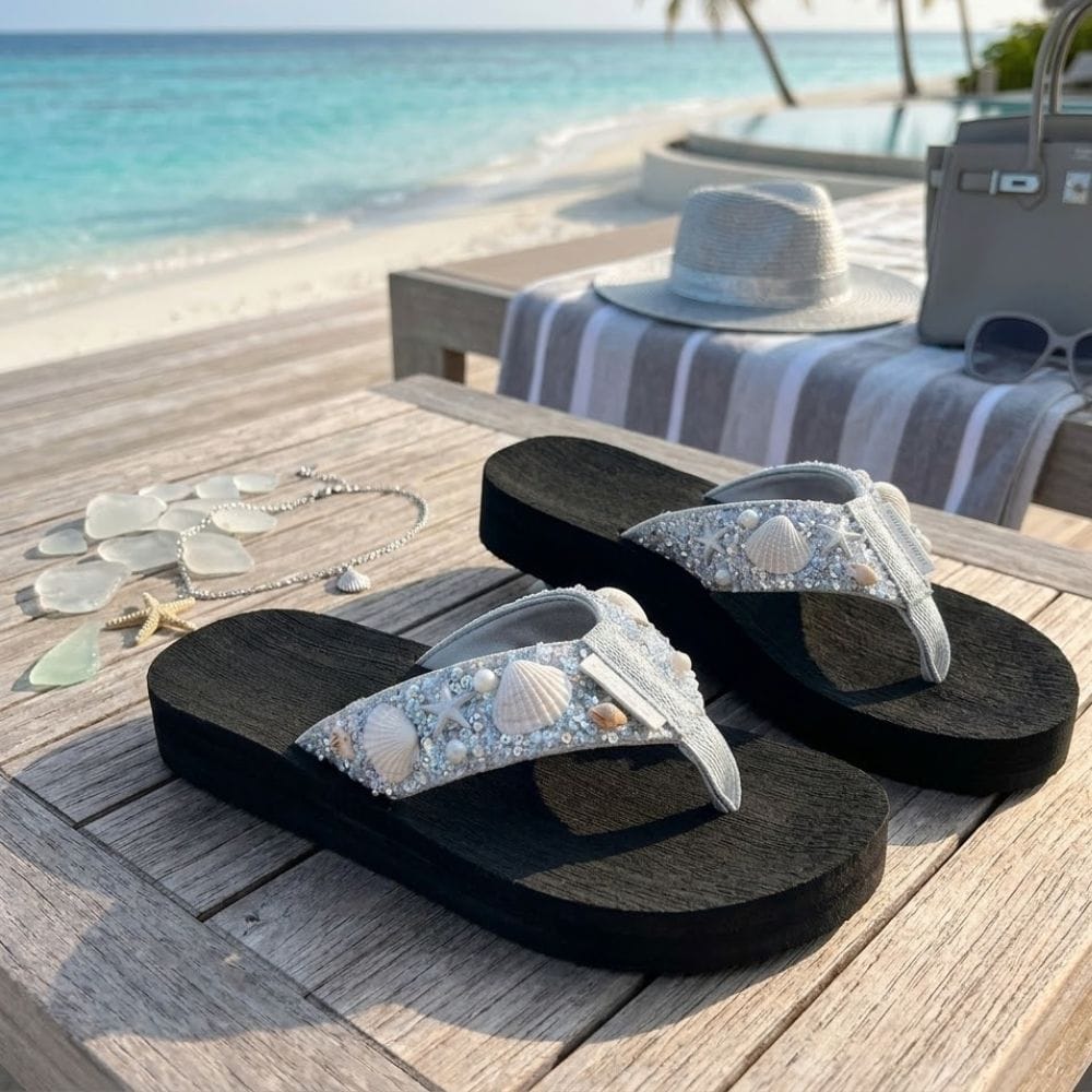 A pair of black flip-flop sandals with embellished silver straps featuring real seashells, displayed on a wooden table at a beach resort with a straw hat, sunglasses, and jewelry nearby.